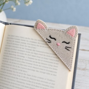May include: A triangular felt corner bookmark shaped like a cat head. The bookmark is light gray with pink inner ears, embroidered black closed eyes, black whiskers, and a small pink stitched nose, placed on the corner of an open book.