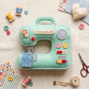 Puede incluir: Un adorno de máquina de coser de fieltro verde menta con el texto bordado STITCH WITH JOY. Presenta un trozo de tela floral azul bajo la aguja, botones de fieltro coloridos, un control de velocidad rojo y una rueda gris. Rodeado de accesorios de costura.