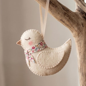 May include: A cream-colored felt bird ornament featuring a small floral print scarf, embroidered closed eyes, and pink cheeks. The bird is suspended by a light beige ribbon, showcasing visible blanket stitching along the edges of its body and wing.