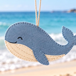 May include: A felt whale ornament in shades of blue and cream, featuring visible stitched edges and a simple smiling face. The whale is suspended by a thin tan string, showcasing a rounded body, a small fin, and a wide tail fluke.