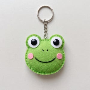 May include: A green felt frog keychain featuring large white eyes with black pupils, pink circular cheeks, and a stitched black smile. The frog head is attached to a silver-colored metal split ring by a short chain, showcasing visible stitching along the edge.