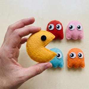 May include: A yellow felt Pac-Man character with a black circular eye, accompanied by four small felt ghost characters in red, pink, light blue, and orange, each featuring large white and black circular eyes and a scalloped bottom edge.