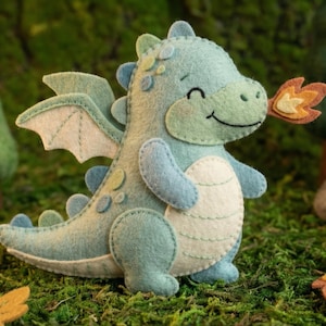 May include: A light blue felt dragon with white accents, wings, and a smiling face. The dragon is exhaling a felt flame. The scene includes felt trees, mushrooms, and leaves, creating a whimsical forest setting.