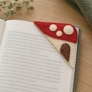 May include: A triangular felt corner bookmark featuring a red mushroom cap top with cream-colored spots and a cream-colored base with a brown stitched door design. The bookmark is placed on the corner of an open book page with visible stitching.