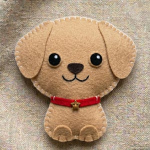 May include: A tan felt dog ornament featuring a red collar with a small gold star charm. The dog has black circular eyes, a dark brown nose, and a stitched mouth. The edges of the felt are joined with visible white blanket stitching throughout.