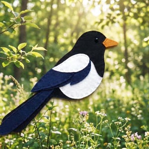 May include: A felt magpie bird with a black head, back, and tail, white wing accents, and an orange beak. The bird is set against a blurred background of a forest with green foliage and sunlight.