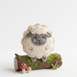 May include: A handcrafted felt sheep with a cream-colored body, gray face, and pink cheeks, perched on a moss-covered log. Two small mushrooms and grass accents are at the base, creating a whimsical woodland scene.
