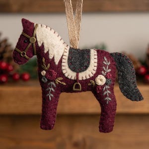May include: A burgundy felt horse ornament with a white mane and floral embroidery. The horse has a grey saddle and tail, and is suspended by a beige ribbon. The ornament is approximately 13 cm long.