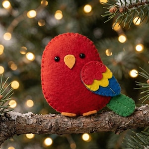 May include: A handmade red felt bird ornament with black eyes, an orange beak, and colorful wing details. The bird is perched on a snowy branch with a Christmas tree and bokeh lights in the background. A festive holiday decoration.