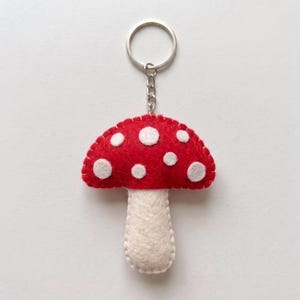 May include: A felt mushroom keyring featuring a bright red cap with white polka dots and a cream-coloured stem. The mushroom is constructed with visible blanket stitching along the edges and is attached to a silver-toned metal split ring.