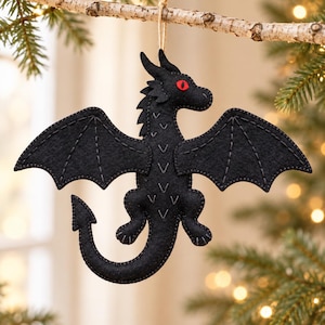 May include: A black felt dragon ornament featuring red eyes and grey stitched detailing on the wings and body. The dragon has a long curved tail and is suspended by a thin twine string, designed for hanging as a decorative piece.