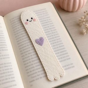 May include: A cream-colored felt bookmark featuring a stitched face with black eyes, a small smile, and pink cheeks. The bookmark has a light purple heart stitched in the center and a scalloped bottom edge, resting on an open book.