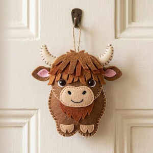 Può includere: Ornamento artigianale in feltro a forma di mucca delle Highlands. La mucca marrone ha una criniera ispida, corna color crema e orecchie rosa. È appesa a uno spago di iuta su una porta bianca. La mucca ha un volto sorridente.