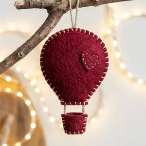 May include: A burgundy felt hot air balloon ornament with a stitched heart detail, suspended from a twig. The balloon has a white stitched outline and a small basket. Decorative lights are in the background.