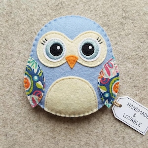 May include: A handmade blue felt owl with large black eyes, an orange beak, and cream-colored accents. The owl has patterned wings with floral designs and a tag that reads "Handmade & Lovable."