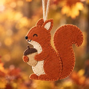 May include: A felt squirrel ornament in shades of rust orange and cream, featuring visible stitched edges and a small brown acorn held in its paws. The squirrel is suspended by a thin twine loop, showcasing a flat, layered construction.