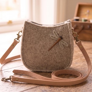 May include: A beige felt crossbody bag with a rounded bottom and a stitched dragonfly appliqué on the front. The bag features a long, light tan leather strap attached with brass-toned metal swivel clasps on both sides of the structured felt body.
