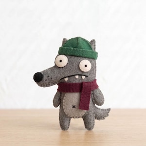 May include: A handcrafted felt wolf figurine in shades of grey, wearing a dark green beanie and a deep maroon scarf. The wolf has large white eyes with black pupils, a black nose, and a light grey belly patch with a small black cross stitch.