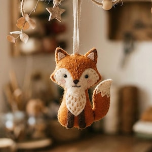 Puede incluir: Adorno de zorro de fieltro hecho a mano con vientre blanco, punta de la cola y detalles. El zorro es naranja y marrón, con una expresión dulce, suspendido por una cuerda. Elementos decorativos: estrella y hojas de madera.