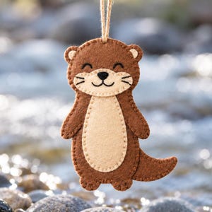 May include: A brown and beige felt otter ornament featuring a stitched face with a black nose and whiskers. The otter has a light-colored belly patch and is suspended by a thin tan twine loop attached to the top of its head.