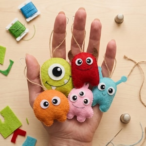 Könnte beinhalten: Fünf kleine Filzmonster-Anhänger in leuchtenden Farben wie Limettengrün, Rot, Orange, Pink und Hellblau. Jedes Monster verfügt über gestickte Details, Wackelaugen und eine Schnurschlaufe zum Aufhängen, mit verschiedenen Formen und Gesichtsausdrücken.