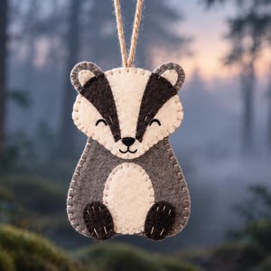 May include: A felt badger ornament featuring a cream and dark gray color scheme with visible whip-stitch detailing along the edges. The badger has a white face with dark gray stripes, closed eyes, and a small black nose, suspended by a twine loop.