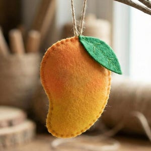 May include: A felt mango ornament with a gradient of yellow to orange hues, and a green leaf. The mango is hanging from a natural twine string. The background is blurred, with wooden items and a spool of twine.