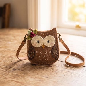 May include: A brown felt owl-shaped crossbody bag featuring cream-colored eye patches, embroidered feather details, and a small decorative cluster of green leaves and red berries attached to the top corner, complete with a thin tan leather strap.