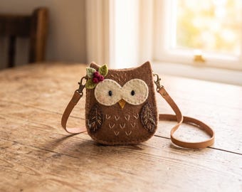 May include: A brown felt owl-shaped crossbody bag featuring cream-colored eye patches, embroidered feather details, and a small decorative cluster of green leaves and red berries attached to the top corner, complete with a thin tan leather strap.