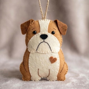 May include: A handmade felt bulldog ornament in shades of brown, white, and tan. The dog has a sad expression and a small heart. It is designed with a string for hanging. Ideal for pet lovers.