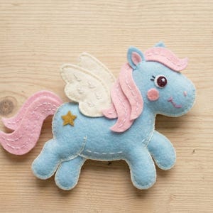 May include: A light blue felt pegasus ornament featuring a pink mane and tail, cream-colored wings with stitched details, and a small gold star appliqué on its flank. The figure has a dark eye, a pink cheek, and white decorative stitching.