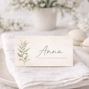Könnte beinhalten: Eine cremefarbene Tischkarte mit dem Namen "Anna" in Schreibschrift, verziert mit einer Aquarell-Olivenzweig-Illustration. Die Karte liegt auf einer weißen Leinen-Serviette, auf einem weißen Teller, passend für eine Hochzeit oder ein Event.