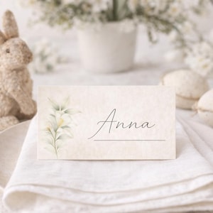 Könnte beinhalten: Eine beige Tischkarte mit dem Namen "Anna" in Schreibschrift, verziert mit einem floralen Design. Die Karte liegt auf einer weißen Leinen-Serviette, mit einem dekorativen Hasen und Osterei-Ornamenten im Hintergrund.