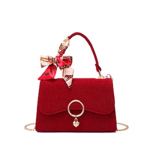 Puede incluir: Un bolso rojo con una correa de cadena dorada y un asa a juego. El bolso presenta un pañuelo decorativo atado al asa y un cierre de anillo dorado con un dije en forma de corazón. El bolso está hecho de un material texturizado.