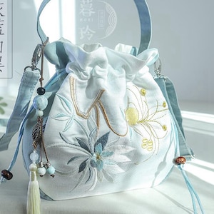 Puede incluir: Un bolso con cordón azul claro con diseños florales bordados en tonos azules, dorados y blancos. El bolso tiene un asa y una correa para el hombro azules a juego, borlas de cuentas decorativas y un cierre de cordón.