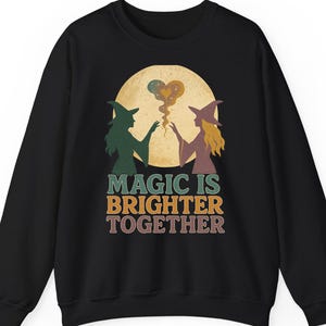 May include: Black sweatshirt with a graphic featuring two witches, a full moon, and the text "MAGIC IS BRIGHTER TOGETHER." The witches are in silhouette, one green and one gold, with a heart-shaped design between them.
