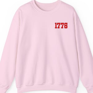 May include: A light pink sweatshirt with the number "1776" printed in red on the upper left chest. The sweatshirt has a crew neck and long sleeves.