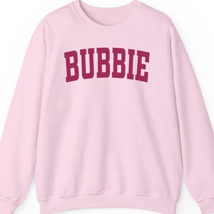 May include: A light pink sweatshirt with the word "BUBBIE" printed in maroon block letters across the chest. The sweatshirt has a crew neck and long sleeves, and is made of a soft material.