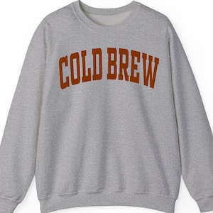 May include: A grey marl sweatshirt featuring the text "COLD BREW" in a burnt orange, arched font. The crew neck sweatshirt has long sleeves, suitable for cooler weather and casual wear.