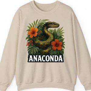 Könnte beinhalten: Beiges Sweatshirt mit detaillierter Illustration einer grünen Anakonda, die sich inmitten üppigen grünen Laubes und leuchtend orangefarbener Blumen windet. Das Wort "ANACONDA" ist in fetten, schwarzen Buchstaben am unteren Rand des Designs aufgedruckt.
