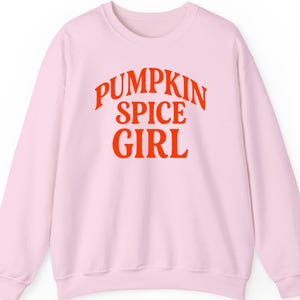 Könnte beinhalten: Hellrosa Sweatshirt mit dem Schriftzug "PUMPKIN SPICE GIRL" in leuchtendem Orange auf der Brust. Das Sweatshirt hat einen Rundhalsausschnitt und lange Ärmel, ideal für kühleres Wetter.