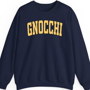 Könnte beinhalten: Marineblaues Sweatshirt mit dem Wort "GNOCCHI" in gelben Blockbuchstaben auf der Brust. Das Sweatshirt hat einen Rundhalsausschnitt und lange Ärmel. Der Stoff scheint weich und bequem zu sein.