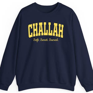 Op de afbeelding: Marineblauwe sweatshirt met het woord "CHALLAH" in gele blokletters. Daaronder staat de zin "Soft. Sweet. Sacred." in kleinere, gele letters. De sweatshirt heeft een ronde hals en lange mouwen.