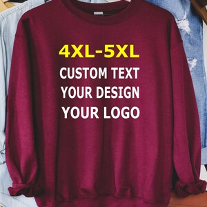 May include: A maroon sweatshirt with the text "4XL-5XL CUSTOM TEXT YOUR DESIGN YOUR LOGO" in white and yellow. The sweatshirt is hanging on a wooden hanger with other clothing items in the background.