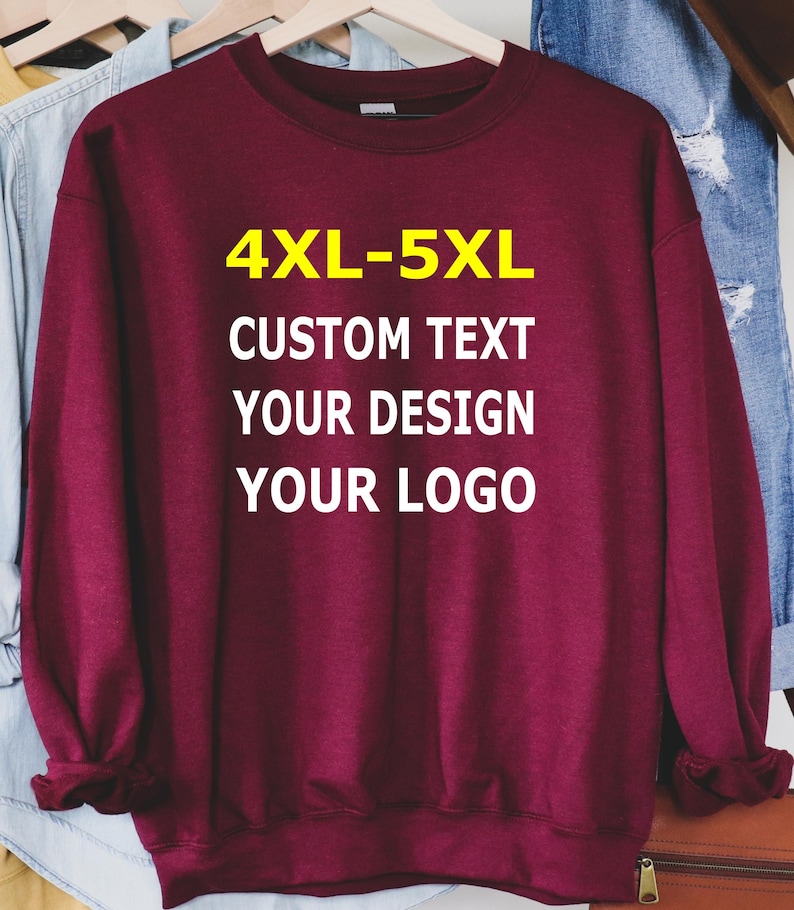 May include: A maroon sweatshirt with the text "4XL-5XL CUSTOM TEXT YOUR DESIGN YOUR LOGO" in white and yellow. The sweatshirt is hanging on a wooden hanger with other clothing items in the background.