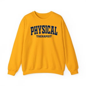 K&ouml;nnte beinhalten: Senfgelbes Sweatshirt mit dem Aufdruck "PHYSICAL THERAPIST" in marineblauen Gro&szlig;buchstaben. Das Sweatshirt hat einen Rundhalsausschnitt und lange &Auml;rmel. Der Text ist mittig auf der Brust platziert.