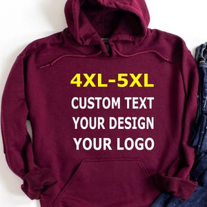 May include: A maroon hooded sweatshirt with a front pocket and drawstring. The text "4XL-5XL CUSTOM TEXT YOUR DESIGN YOUR LOGO" is printed in yellow and white on the front. A pair of blue jeans is visible on the right side.