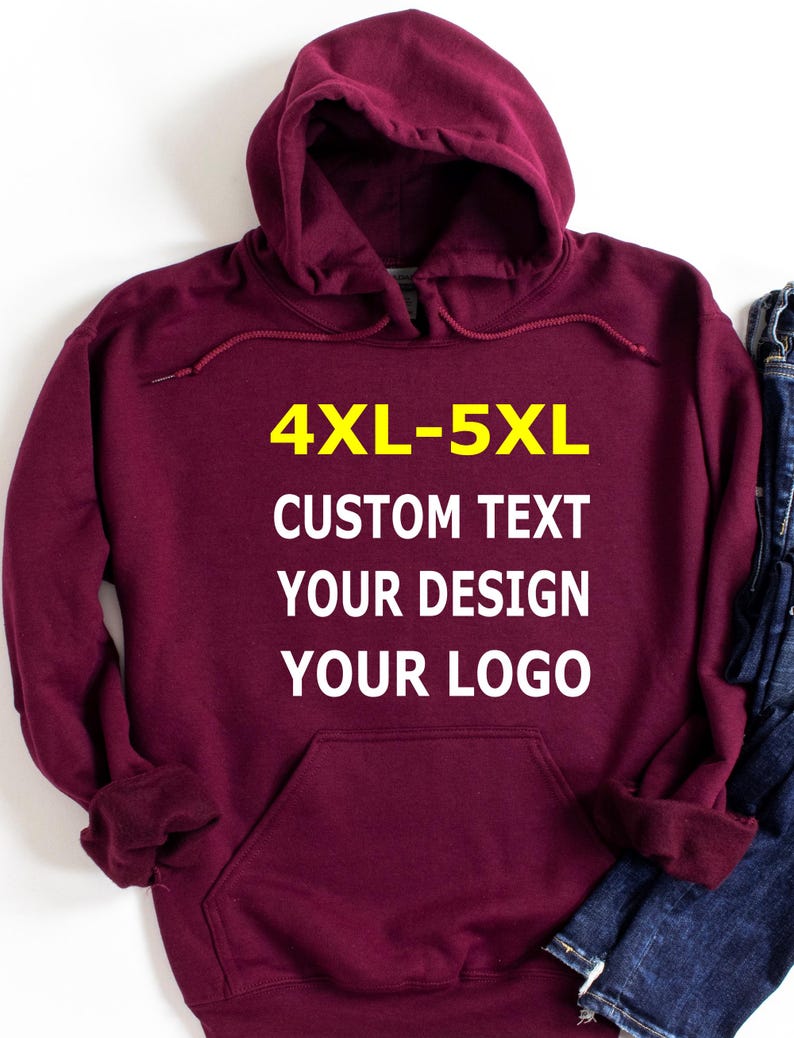 May include: A maroon hooded sweatshirt with a front pocket and drawstring. The text "4XL-5XL CUSTOM TEXT YOUR DESIGN YOUR LOGO" is printed in yellow and white on the front. A pair of blue jeans is visible on the right side.
