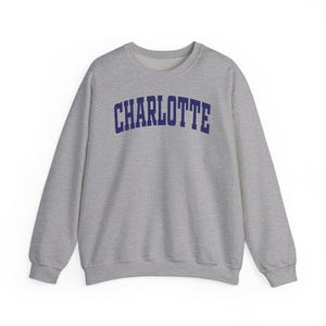 May include: A heather gray sweatshirt with the word "CHARLOTTE" printed across the chest in navy blue, arched lettering. The crew neck sweatshirt has long sleeves and a relaxed fit, suitable for casual wear.