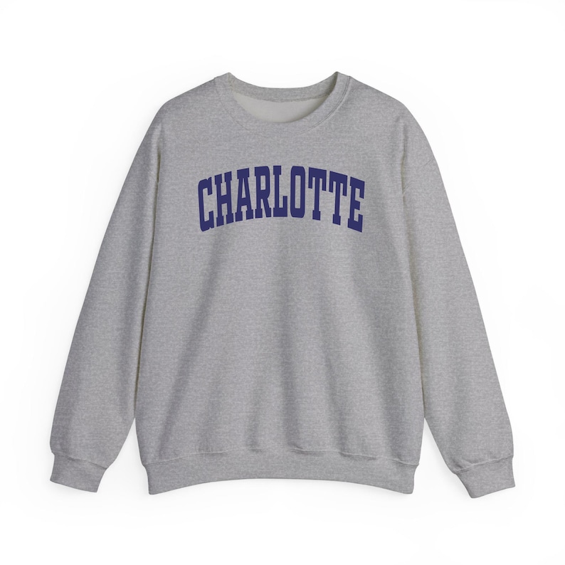 May include: A heather gray sweatshirt with the word "CHARLOTTE" printed across the chest in navy blue, arched lettering. The crew neck sweatshirt has long sleeves and a relaxed fit, suitable for casual wear.